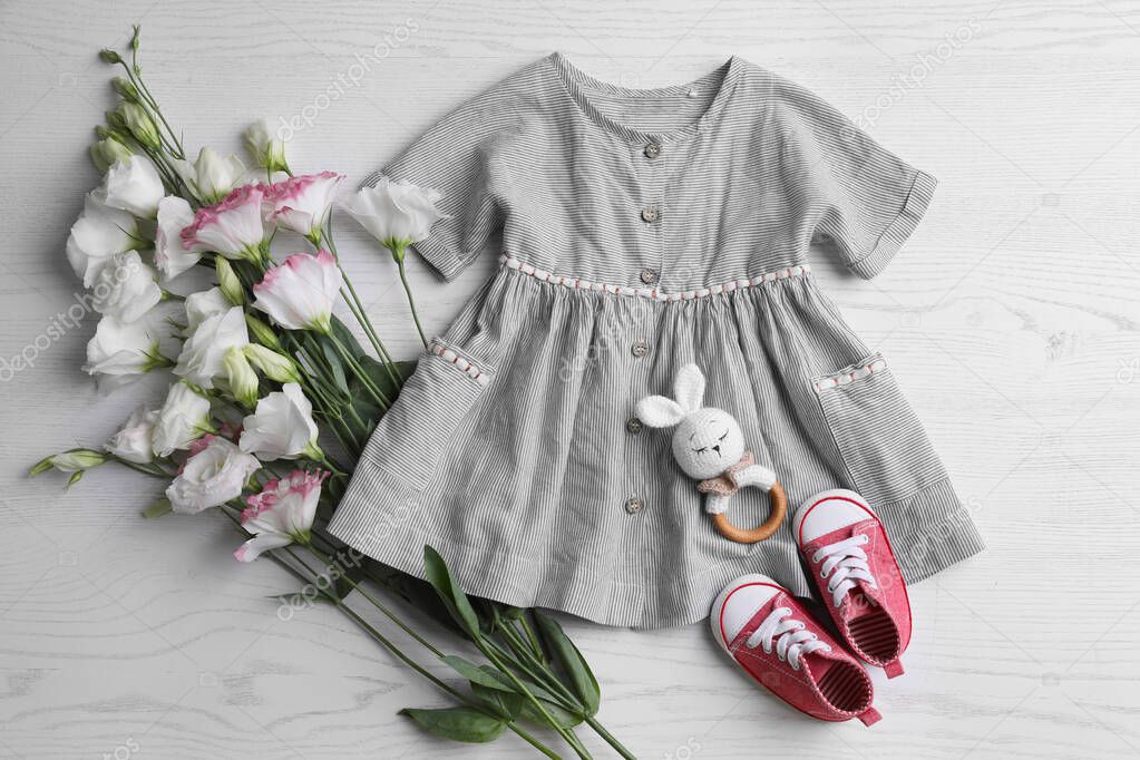 Baby dress, shoes, toy and flowers on white wooden table, flat lay