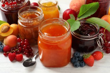 Farklı reçelli kavanozlar ve beyaz ahşap masada taze meyveler.