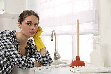 Unhappy young woman with plunger near clogged sink in kitchen clipart