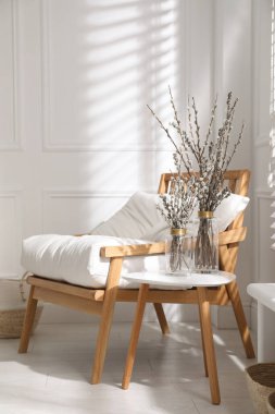 Glass vases with pussy willow tree branches on table near armchair in room