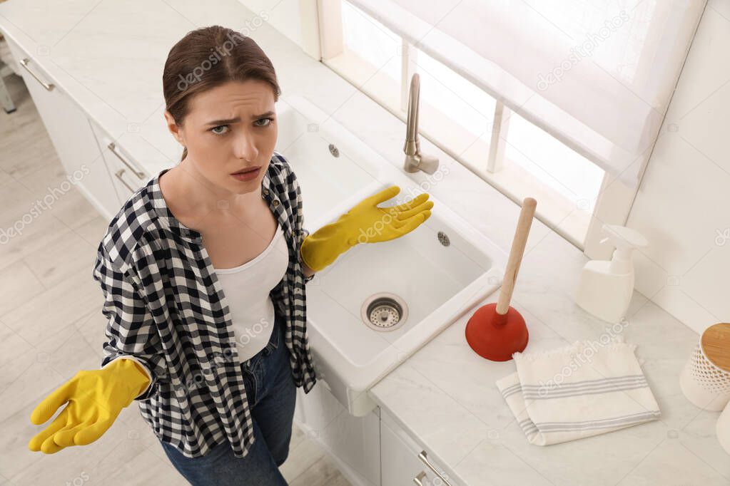 Upset young woman with plunger near sink in kitchen, above view