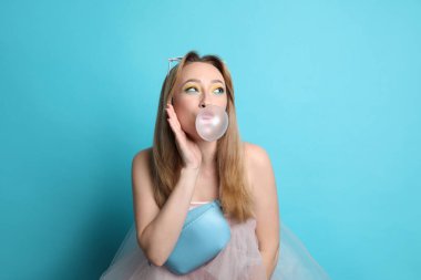 Fashionable young woman with bright makeup blowing bubblegum on light blue background clipart