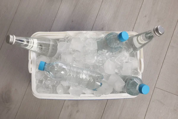 Plastic cool box with ice cubes and bottles of water on wooden floor, top view
