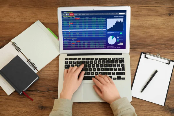 Woman analyzing electronic trading platform on laptop at wooden table, above view. Stock exchange