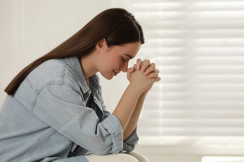 Religious young woman with clasped hands praying indoors