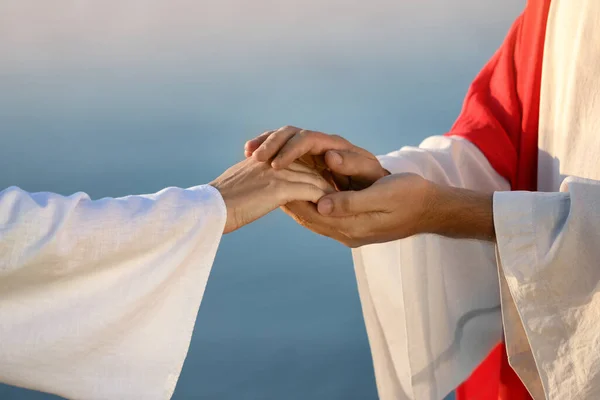 Jesus Christ holding woman's hand near water outdoors, closeup