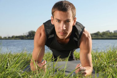 Sportif adam nehir kenarındaki yeşil çimlerde kalas egzersizi yapıyor.