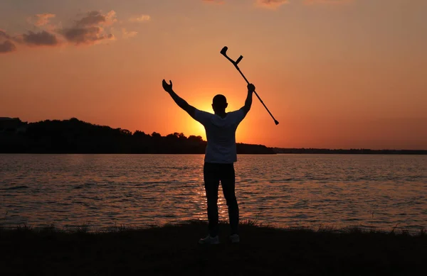 日落时分 男人把肘部的拐杖举到河边的天空中 回头看 治疗奇迹 — 图库照片