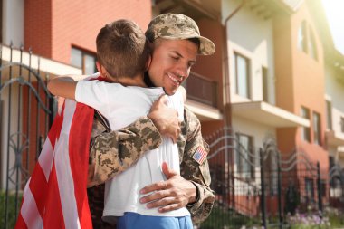 Amerikan bayrağı taşıyan bir asker ve küçük oğlu dışarıda sarılıyor.