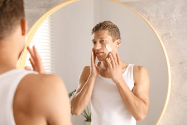 Yakışıklı genç adam banyoda aynanın yanında yüzüne krem sürüyor.