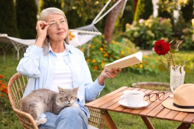 Bahçedeki ahşap masada kedili kitap okuyan yaşlı bir kadın.