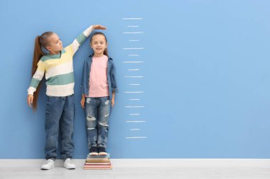 Küçük kız arkadaşının boyunu ölçüyor. Mavi duvarın yanında. Kapalı alanda.