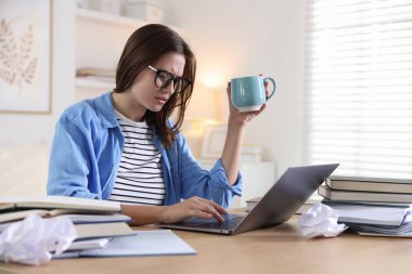 Gözlüklü, dizüstü bilgisayarlı, kitaplı ve fincanlı endişeli öğrenci kapalı bir masada sınava hazırlanıyor.