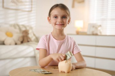 Küçük kız, ahşap masadaki domuz kumbarasına cep harçlığı koyuyor.