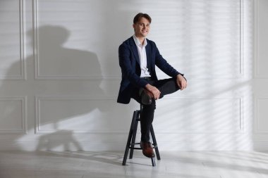 Beyaz duvarın yanındaki taburede şık kotlu bir adam.