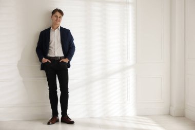 Beyaz duvarın yanında şık kotlu bir adam.