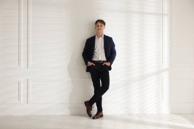 Beyaz duvarın yanında şık kotlu bir adam.