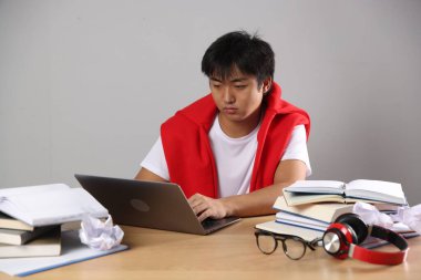 Dizüstü bilgisayarı ve kitapları olan öğrenci gri duvarın yanındaki tahta masada sınava hazırlanıyor.