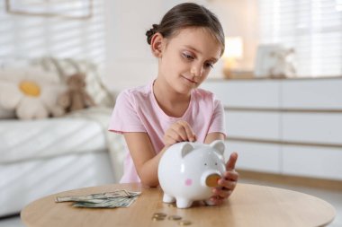 Küçük kız, ahşap masadaki domuz kumbarasına cep harçlığı koyuyor.