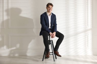 Beyaz duvarın yanındaki taburede şık kotlu bir adam.