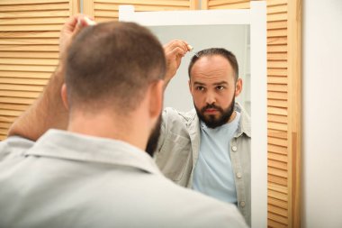 Kellik tedavisi. Evdeki aynanın yanındaki saçına serum süren bir adam.