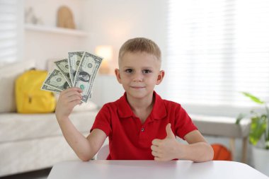 Küçük çocuk cep harçlığı tutuyor ve beyaz masada baş parmak gösteriyor.