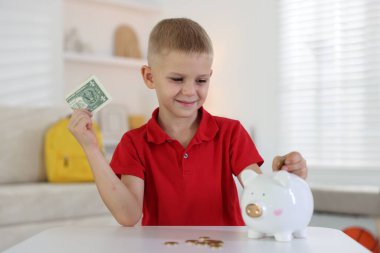 Küçük çocuk, içerideki beyaz masadaki kumbaraya cep harçlığı koyuyor.