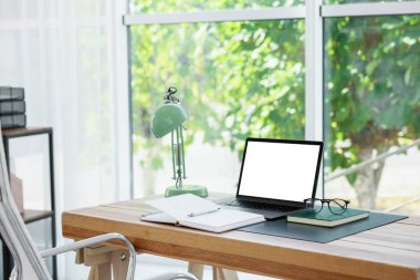 Boş beyaz ekranlı dizüstü bilgisayar, odada lamba ve kırtasiye malzemesi. Rahat ev işyeri