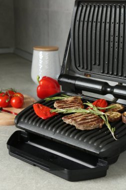 Açık gri masada biftek, biberiye, sebze ve baharatla elektrikli ızgara, yakın plan.