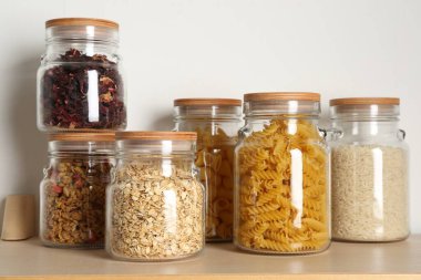 Beyaz duvarın yanındaki ahşap masada farklı ürünler bulunan cam kavanozlar.