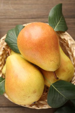 Many fresh pears with green leaves in wicker basket on wooden table, top view