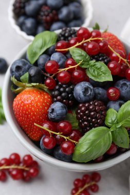 Different ripe berries and basil leaves in bowl on light grey table, closeup