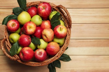 Hasır sepetteki taze elmalar ve ahşap masada yeşil yapraklar, üst manzara. Metin için boşluk