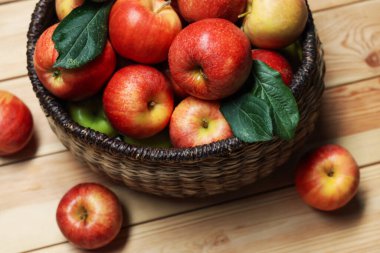 Hasır sepetteki taze elmalar ve yukarıdaki tahta masada yeşil yapraklar.