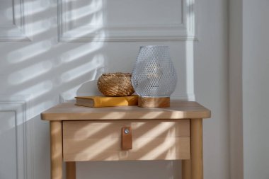 Şık bir gece lambası, hasır depolama kutusu ve beyaz duvarın yanındaki komodinin üzerindeki kitap.