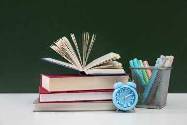 Öğretmenler günün kutlu olsun. Kara tahtaya karşı beyaz masadaki kitaplar, çalar saat ve kırtasiye