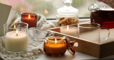 Kadın bardağa sıcak çay dolduruyor ve pencere pervazında mum yakıyor. Sonbahar atmosferi