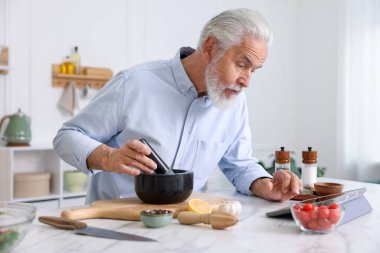 Mutfaktaki beyaz mermer masada yemek pişirirken tablet kullanan yaşlı adam.
