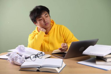 Laptoplu ve kitaplı öğrenci yeşil duvarın yanındaki tahta masada sınava hazırlanıyor.