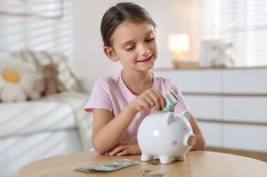 Küçük kız, ahşap masadaki domuz kumbarasına cep harçlığı koyuyor.