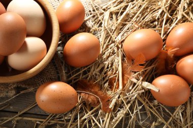 Raw chicken eggs in bowl, feathers and straw on wooden table, flat lay