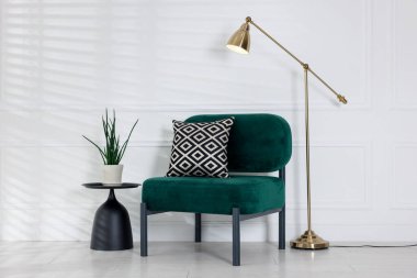 Stylish lamp, comfortable armchair with cushion and houseplant near white wall indoors. Interior design