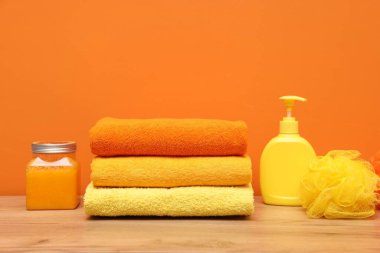 Stack of fresh towels, sponge and cosmetic products on wooden table against orange background