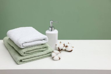 Stack of fresh towels, cotton flowers and cosmetic product on white table against green background, space for text