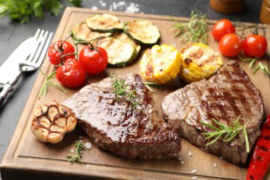 Delicious grilled beef steaks with herbs and vegetables on dark table, closeup