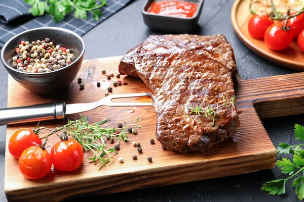Delicious grilled beef steak with herbs, peppercorns, tomatoes, carving fork and ketchup on dark textured table, closeup