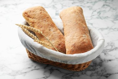 Fresh crispy ciabattas and wheat ears in basket on white marble table, closeup