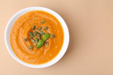 Delicious pumpkin soup with seeds and basil in bowl on beige table, top view. Space for text