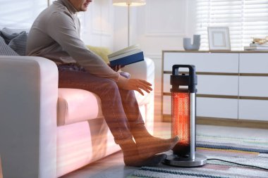 Evinde elektrikli ısıtıcının yanında kitabı olan genç bir adam. Aygıttan çıkan turuncu sıcaklık akışı