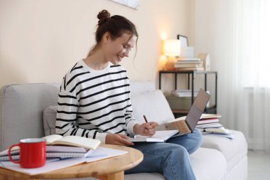 Bilgisayarlı, defterli ve kalemli gülümseyen öğrenci evdeki kanepede sınava hazırlanıyor.